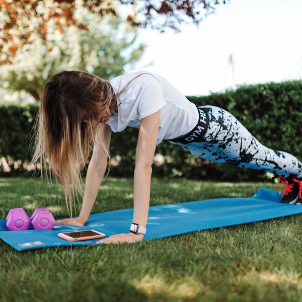 Woman doing push-ups on a yoga mat outside with dumbbells and a smartphone nearby.